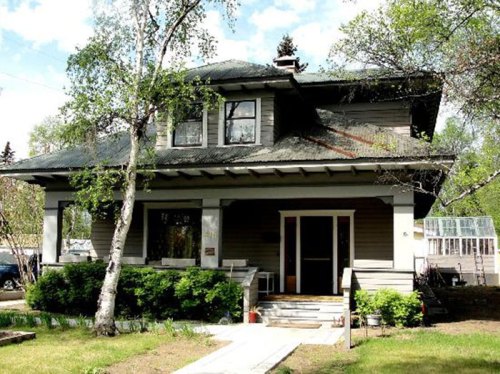 A two-story house with a green roof, surrounded by trees and a well-kept lawn, featuring a front porch and steps.