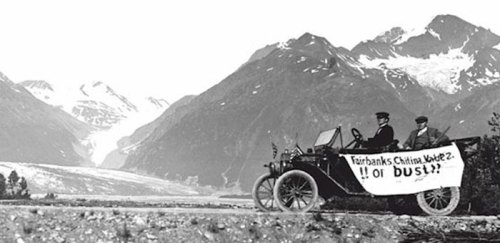 Two men in an early car with a banner reading "Fairbanks Chitina Route 2!! Or Bust!?" against a mountainous backdrop.