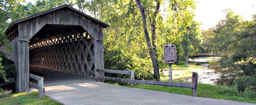 A wooden covered bridge surrounded by lush greenery and a peaceful stream, with a historical marker nearby.