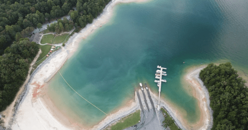 Aerial view of a calm bay with a small marina, surrounded by lush greenery and sandy shores.