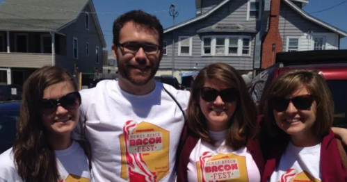 Four people wearing matching T-shirts at a bacon festival, smiling and posing outdoors on a sunny day.