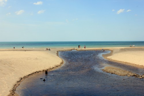 A serene beach scene with a winding stream flowing into the ocean, people walking along the shore under a clear blue sky.