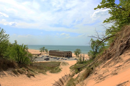 A sandy dune leads down to a beach with a historic building and parked cars near the water under a blue sky.