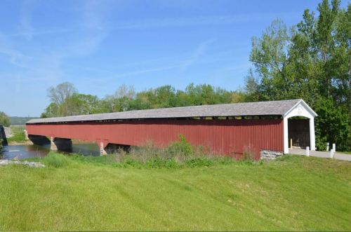 A long red covered bridge spans a river, surrounded by green trees and grass under a clear blue sky.