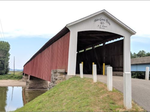 A red covered bridge with a white roof spans a calm waterway, surrounded by green grass and trees.