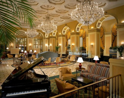 Elegant hotel lobby featuring chandeliers, plush seating, a grand piano, and decorative plants. Warm, inviting atmosphere.
