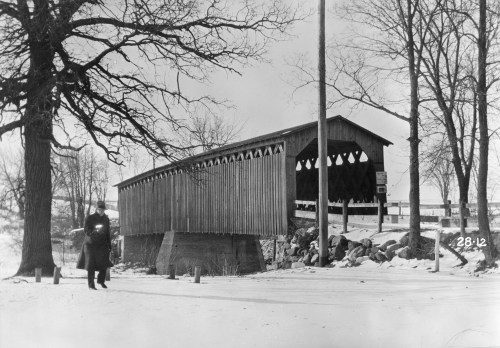 A historic covered bridge stands over a snowy landscape, with a person walking nearby among bare trees.
