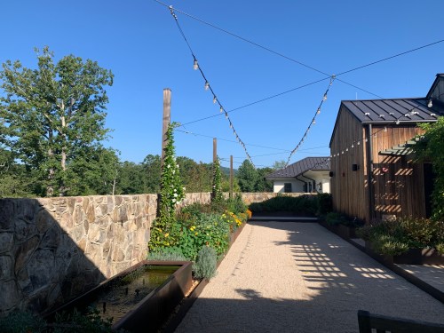 A sunny outdoor space with string lights, stone walls, and lush greenery along a gravel path.