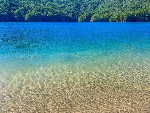 A serene lake with clear blue water and a sandy shore, surrounded by lush green trees and mountains in the background.