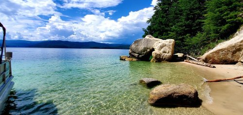 A serene beach scene with clear water, rocky shore, and lush greenery under a blue sky with fluffy clouds.