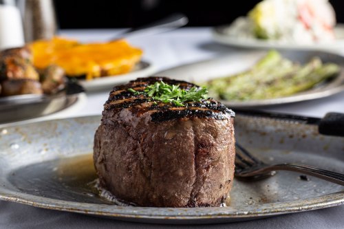 A perfectly grilled steak on a plate, garnished with herbs, with sides of asparagus and cheesy potatoes in the background.