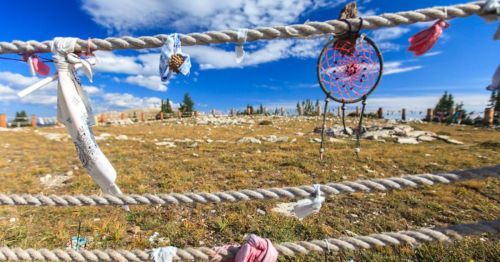 A rope fence adorned with colorful fabric strips and a dreamcatcher, set against a blue sky and grassy landscape.