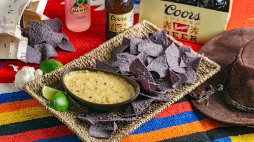 A woven basket holds a bowl of queso dip surrounded by blue corn chips, limes, and drinks on a colorful blanket.