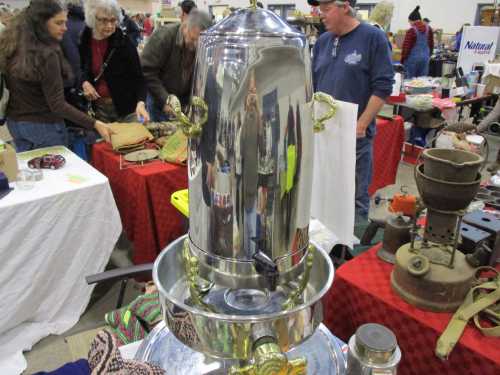 A shiny metal samovar sits on a table, reflecting people and various items at a bustling market or fair.