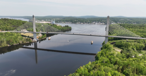 Aerial view of a cable-stayed bridge spanning a river, surrounded by lush greenery and a small town in the background.