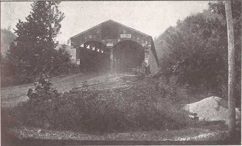 A vintage photograph of a covered bridge surrounded by trees and a gravel path, with a misty atmosphere.