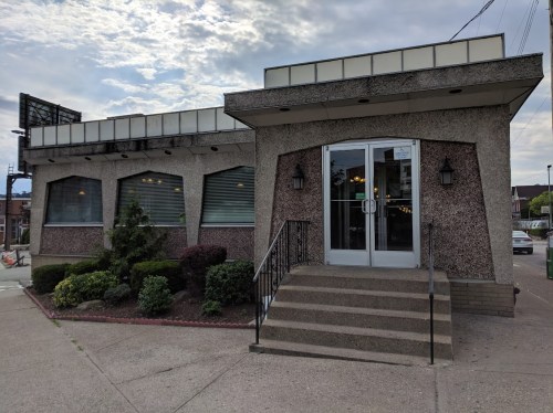 A modern restaurant exterior with large windows, a concrete facade, and steps leading to the entrance.