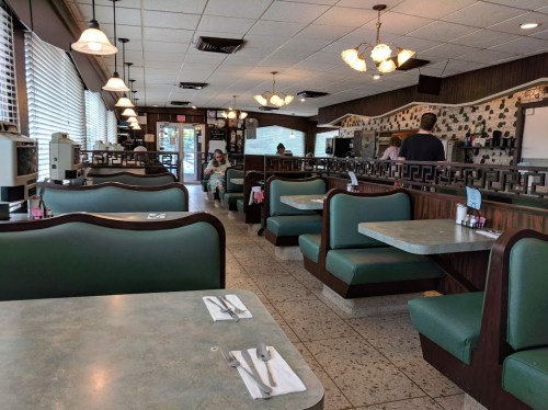 A cozy diner interior with green booths, tables, and a counter, featuring large windows and a relaxed atmosphere.