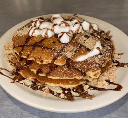 A stack of pancakes topped with whipped cream, chocolate drizzle, and crushed graham crackers on a white plate.