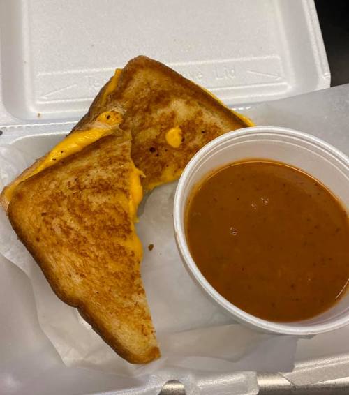 A grilled cheese sandwich with melted cheese, served with a cup of tomato soup in a takeout container.