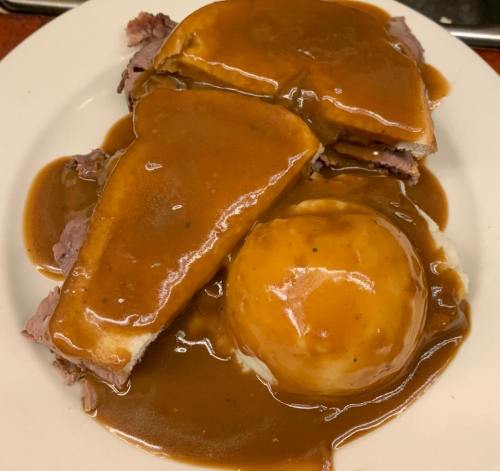A plate of sliced roast beef with gravy, served with bread and a side of mashed potatoes topped with brown gravy.