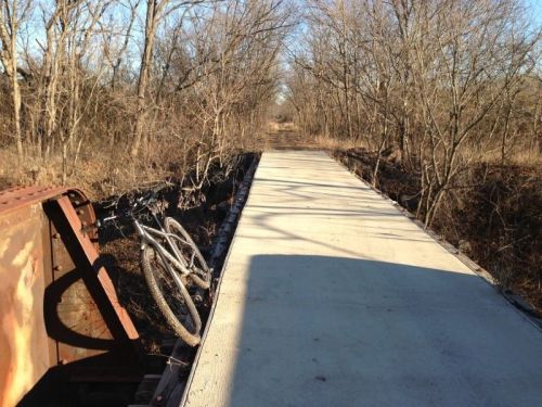 A concrete path through a wooded area, with a bicycle leaning against a rusted metal structure on the side.