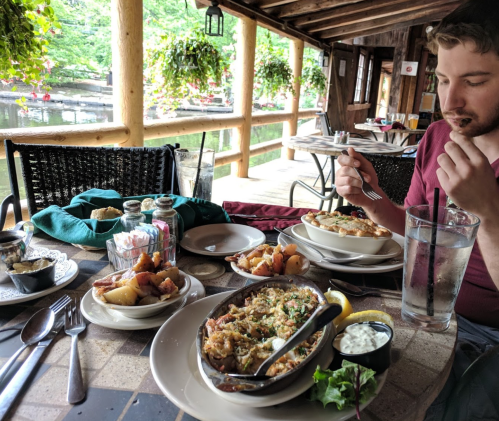 A man enjoys a meal at a rustic outdoor restaurant by the water, with various dishes and drinks on the table.