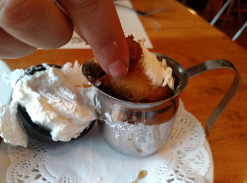 A hand dips a fried dessert into a small metal cup filled with whipped cream, placed on a lace doily.