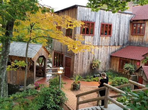 A photographer captures a rustic wooden building surrounded by autumn foliage and colorful flowers.