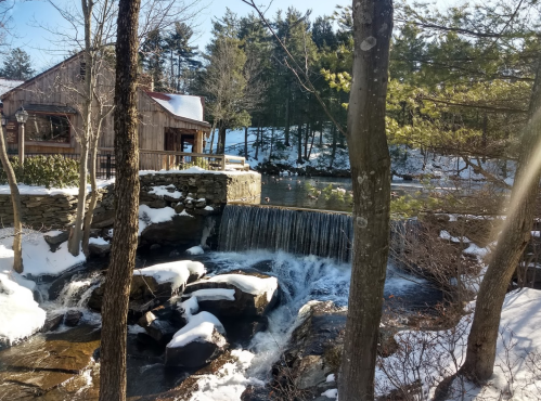 A serene winter scene featuring a small waterfall, rocks, and a rustic wooden building surrounded by snow-covered trees.