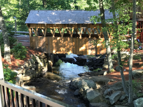 A wooden covered bridge spans a small stream, surrounded by trees and rocky banks in a serene outdoor setting.