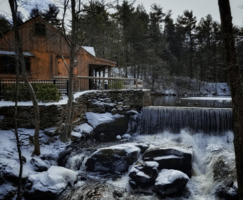A wooden cabin by a snowy river with a waterfall cascading over rocks, surrounded by trees.