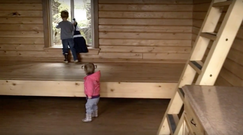 Two children in a wooden room; one looks out a window while the other stands nearby, both dressed for play.