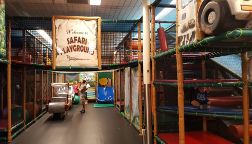 Colorful indoor playground with slides, climbing structures, and children playing. A sign reads "Welcome to Safari Playgrounds."