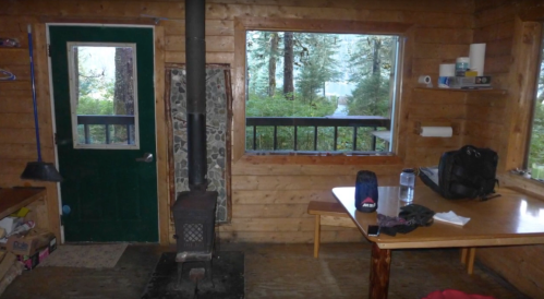Interior of a rustic cabin featuring a wood stove, green door, table, and windows with a view of trees outside.
