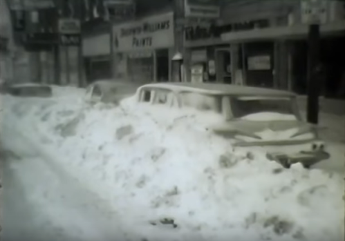 Historic black-and-white photo of cars buried in deep snow on a city street, with storefronts visible in the background.