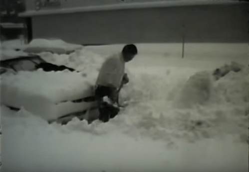 A person shovels snow around a car buried in deep snow during a winter storm.