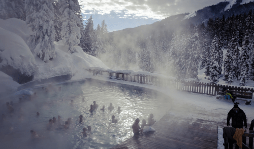 A steaming outdoor hot spring surrounded by snow-covered trees and mountains, with people enjoying the warm water.