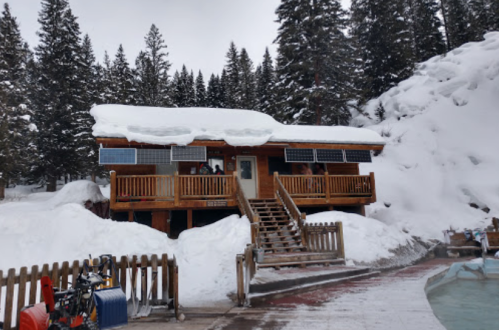 A wooden cabin with solar panels, surrounded by snow and pine trees, featuring a staircase and a wooden fence.