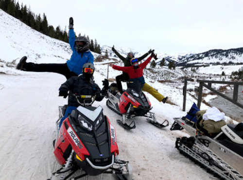 Four people on snowmobiles in a snowy landscape, two striking playful poses, with mountains in the background.