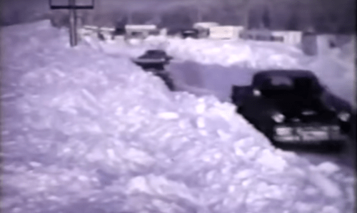 Two cars navigate a snow-covered road, flanked by high snowdrifts in a winter landscape.