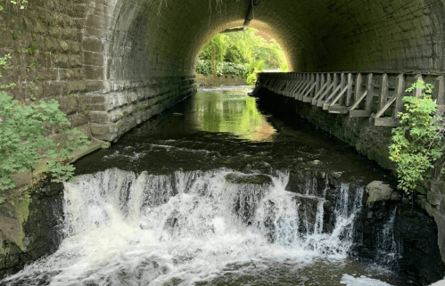 A serene view of a waterway flowing under a stone arch bridge, with greenery on the banks and a small waterfall.