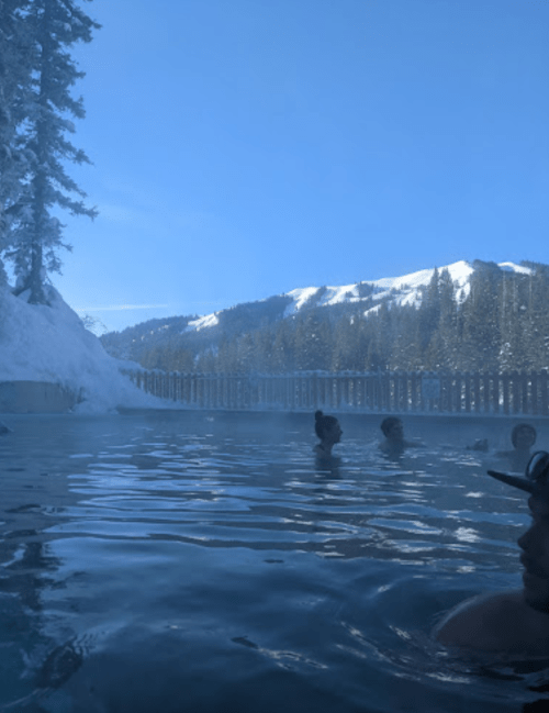 A serene hot spring scene with people relaxing in the water, surrounded by snowy mountains and a clear blue sky.