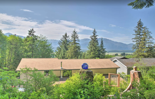 A scenic view of a house surrounded by trees, mountains in the background, and a blue decorative sphere on a stump.