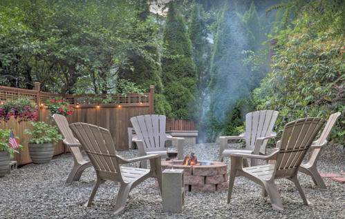 A cozy outdoor fire pit area with Adirondack chairs surrounded by greenery and soft smoke rising in the background.