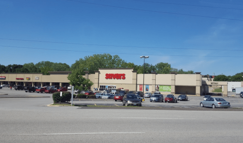 A shopping plaza with a Savers store, surrounded by parked cars and greenery under a clear blue sky.