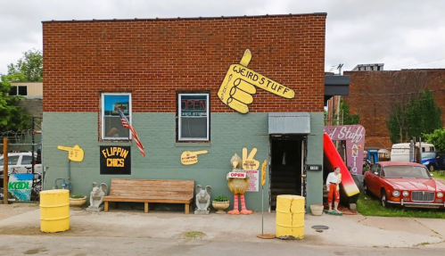 A quirky storefront with colorful signs, a wooden bench, and unique decorations, including a giant chicken and a vintage car.