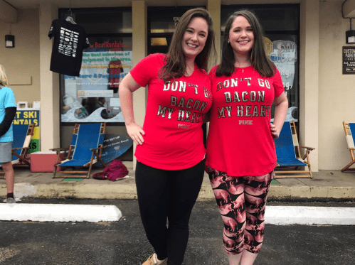 Two women in matching red shirts that say "Don't Go Bacon My Heart," smiling and posing outdoors.
