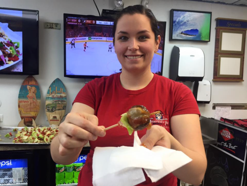 A smiling woman in a red shirt holds a meatball on a stick, with food and a TV in the background.