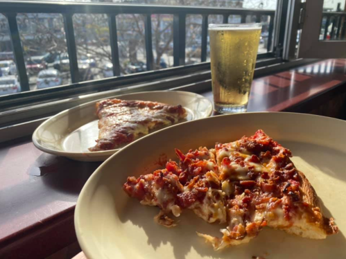 A slice of pizza on a plate next to another slice and a glass of beer, with a view of a parking lot outside.